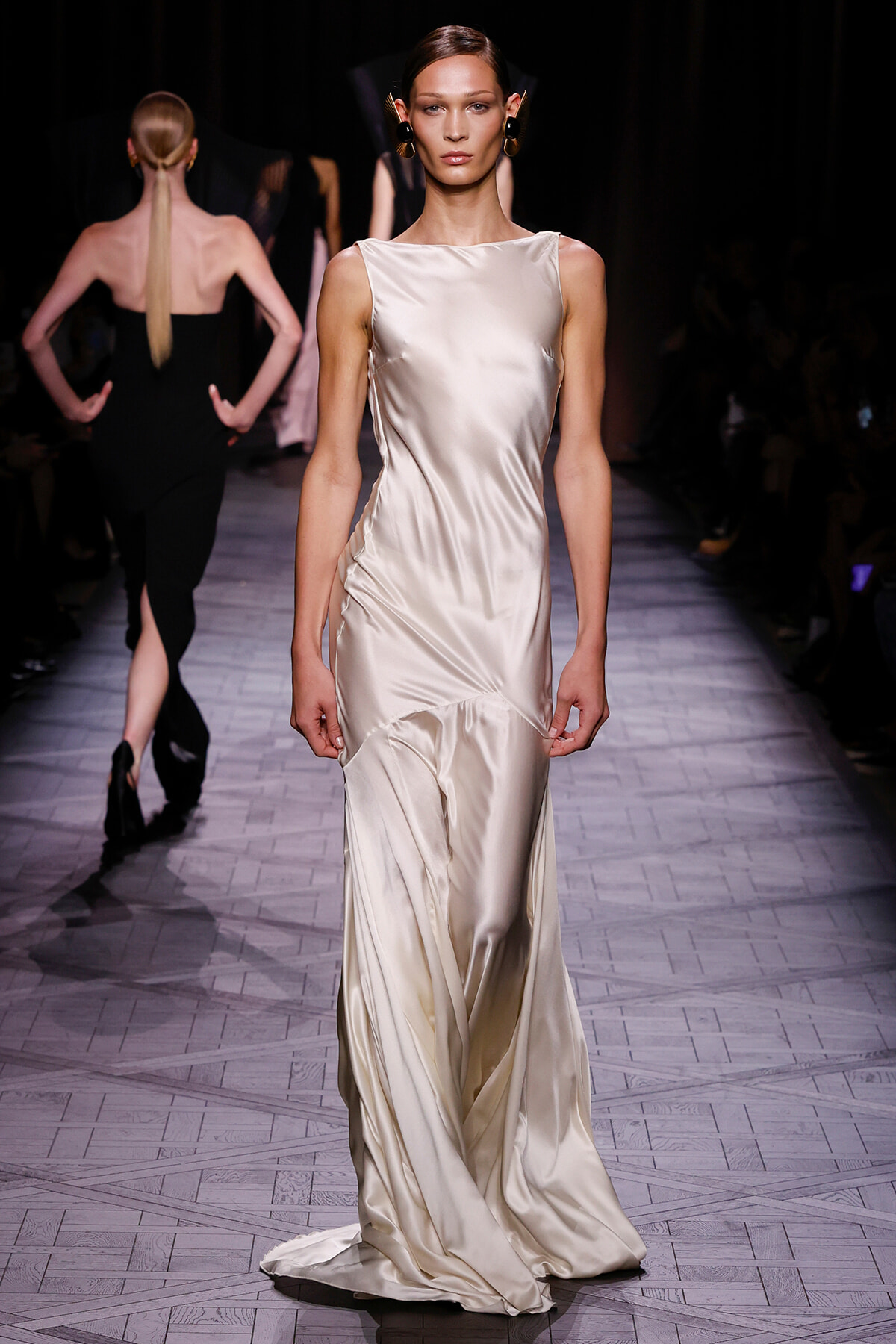 Model walks the runway in a sleeveless ivory satin gown with a fitted bodice and flowing, draped skirt.