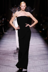 Model on a runway wearing a black strapless gown with a sculptural feathered shoulder detail.
