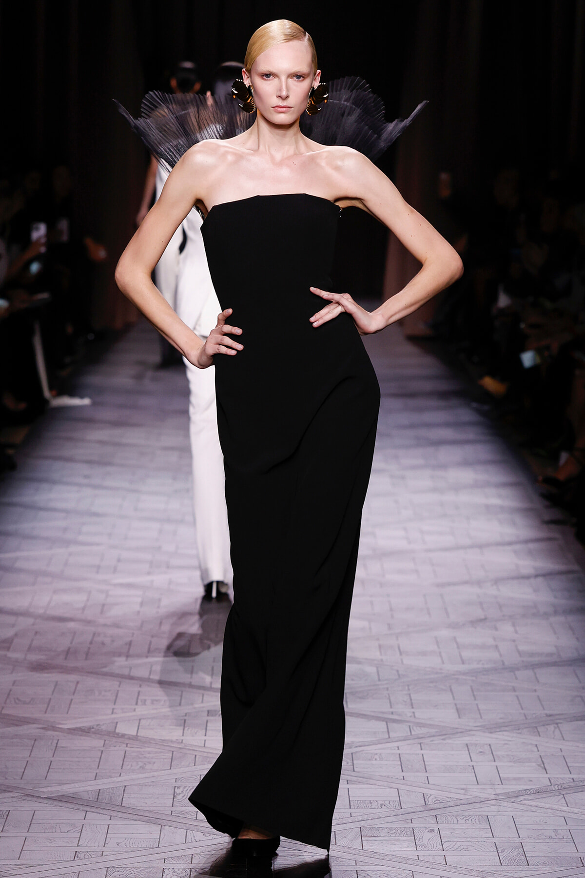 Model on a runway wearing a black strapless gown with a sculptural feathered shoulder detail.