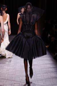 Model on runway wearing a dramatic black pleated gown with a structured corset and oversized sheer collar/neckpiece.