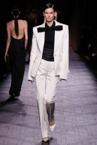 Model walks the runway in a white tailored suit with an oversized blazer, black shirt, and large black bow, paired with slim white trousers and white pointed heels.
