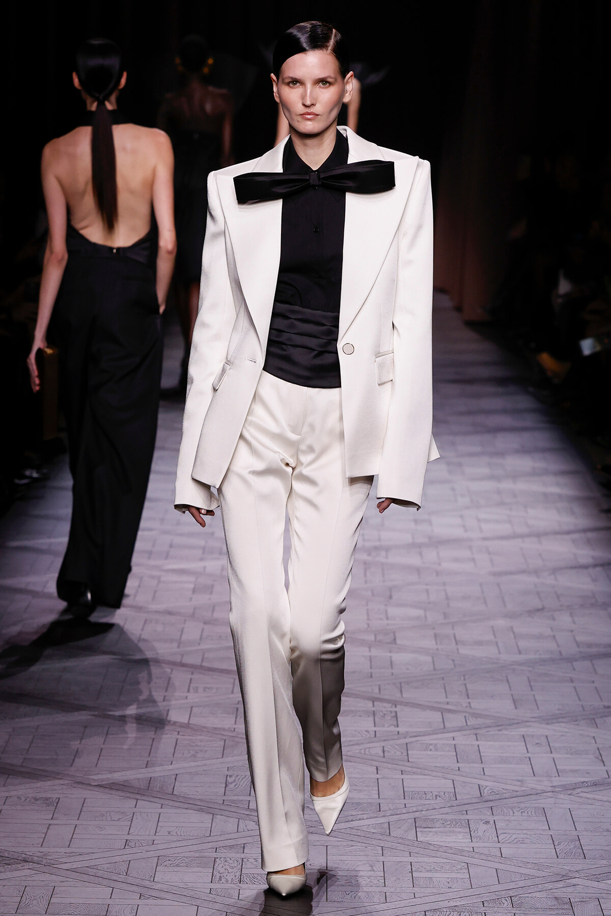 Model walks the runway in a white tailored suit with an oversized blazer, black shirt, and large black bow, paired with slim white trousers and white pointed heels.