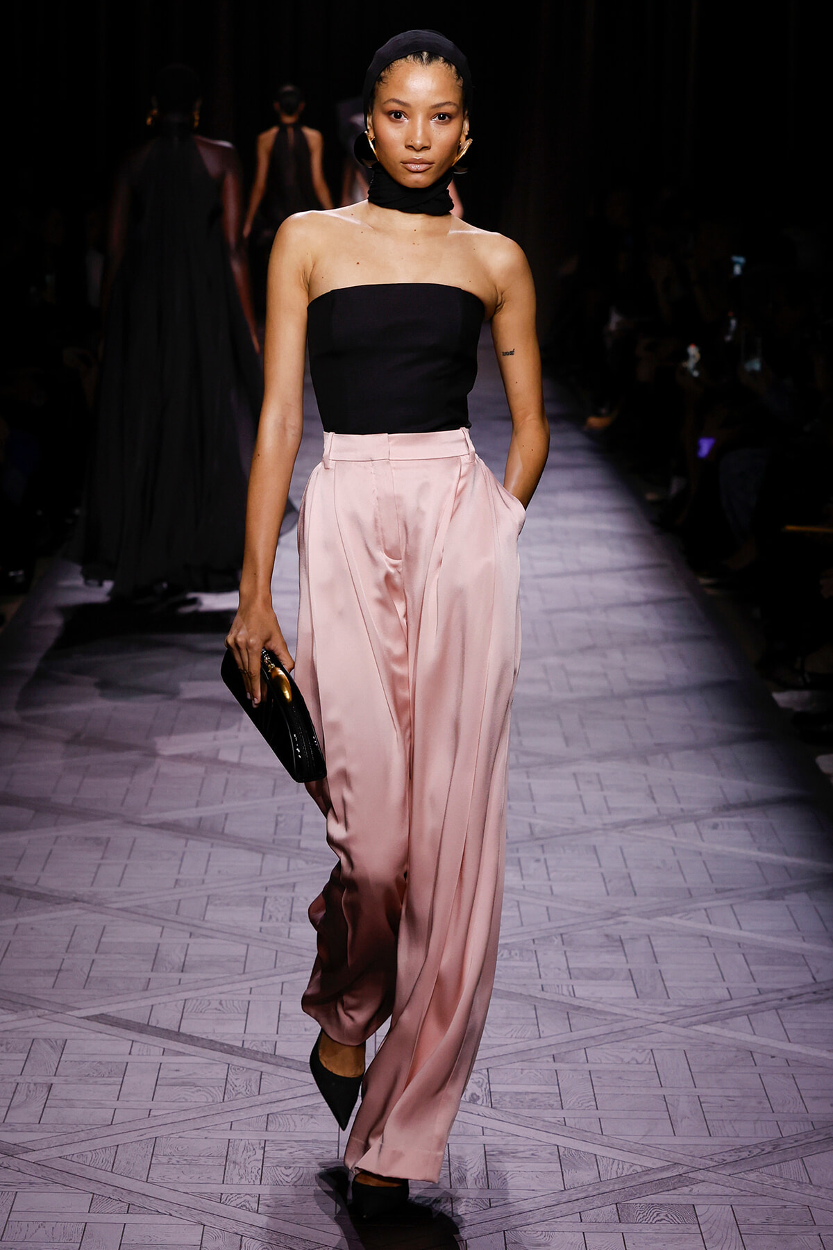 Model walks the runway in a strapless black top and pale pink satin wide-leg pants, carrying a small black clutch.