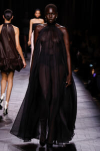 Model walking a fashion show in a floor-length, sheer black halter gown with a flowing silhouette on the runway.