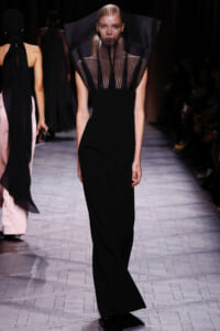 Model walking the runway in a black, floor-length gown with a sheer pleated cape-like neckline.