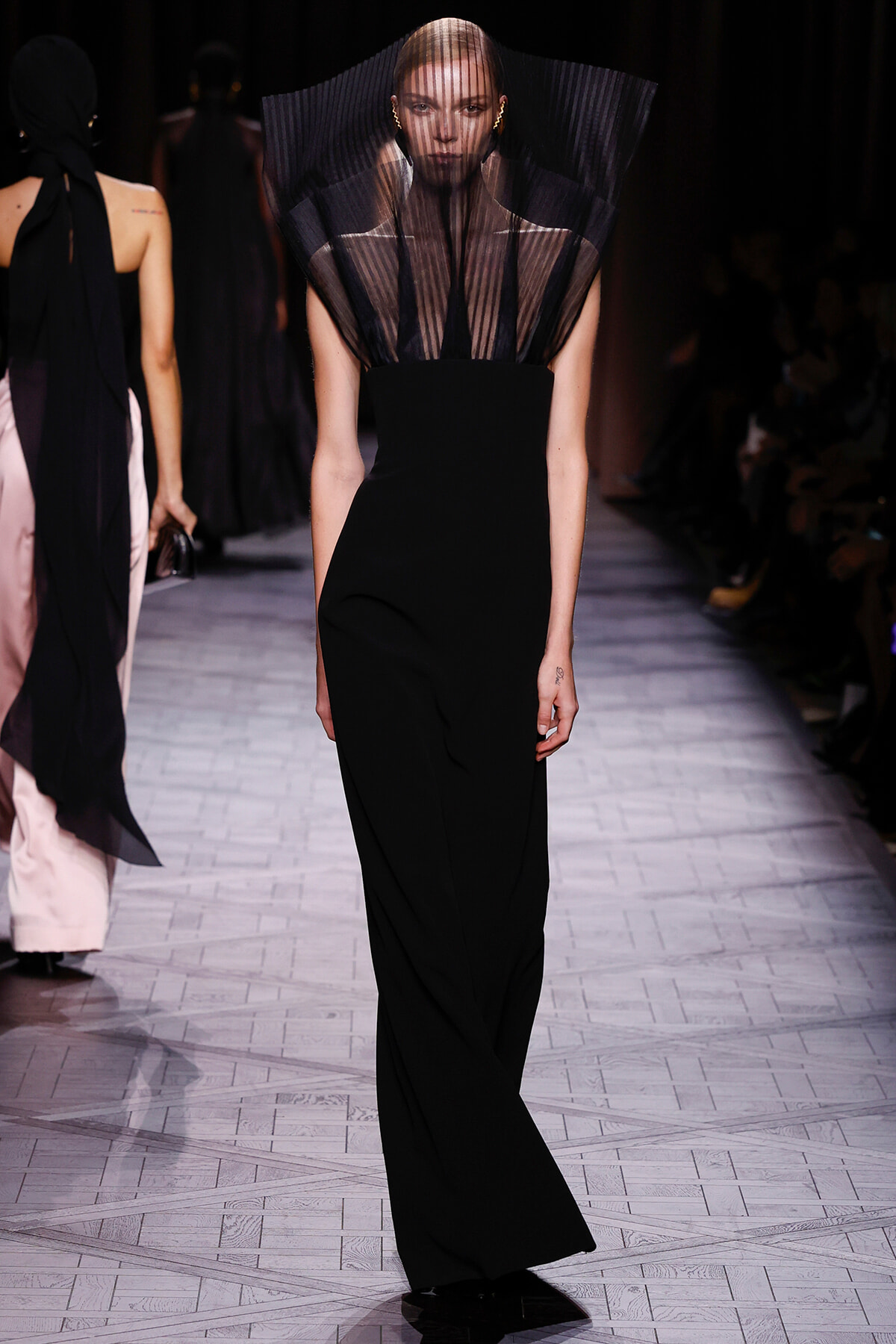 Model walking the runway in a black, floor-length gown with a sheer pleated cape-like neckline.