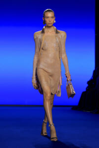Model walking a runway in a gold shimmering knit dress with matching cardigan, carrying a small metallic handbag against a blue gradient backdrop.