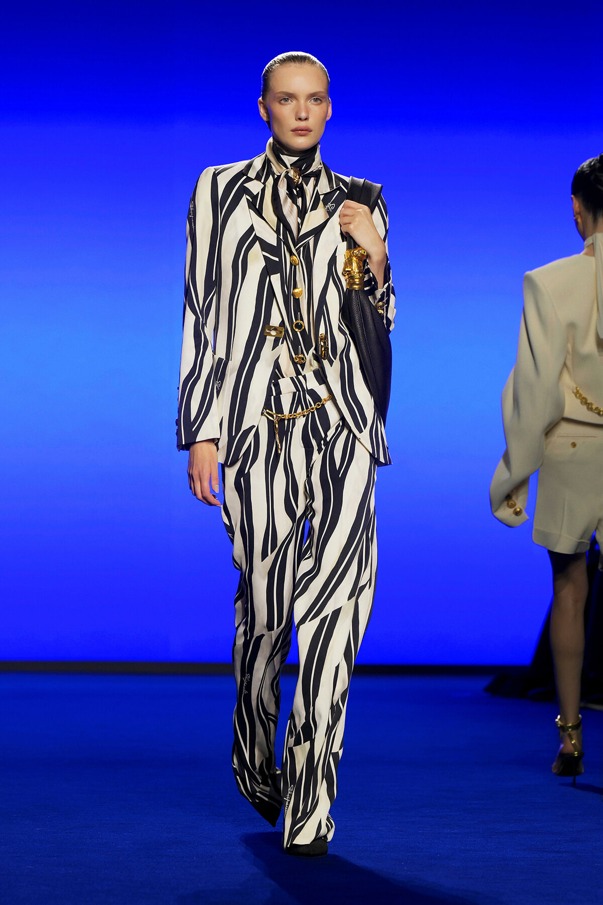Model walks the runway in a bold black-and-white zebra-striped suit with a high-neck scarf, accessorized with gold jewelry and a black handbag against a blue backdrop.