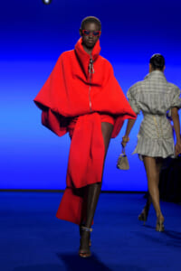 Model on a runway wearing a bright red oversized cape coat with a high collar and matching red shorts, red sunglasses, walking toward the camera against a blue backdrop; a second model in a gray plaid outfit appears in the background.