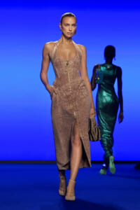 Model on a runway wearing a fitted beige velvet corset dress with a front gold zipper and slit, against a blue backdrop.