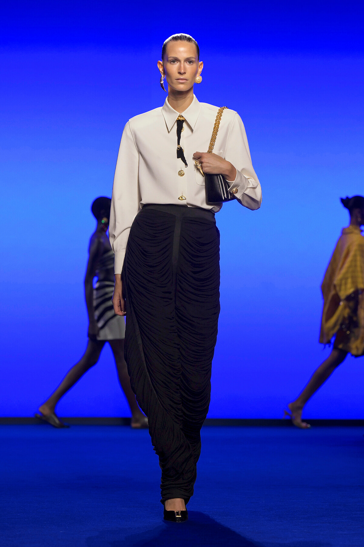 Model walking a runway in a white blouse with gold buttons and a black ruched skirt, carrying a black bag with a gold chain against a blue backdrop.