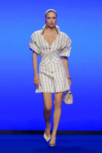 Model walks the runway in a white grid-pattern dress with gold buttons and dramatic rolled sleeves, against a blue backdrop.