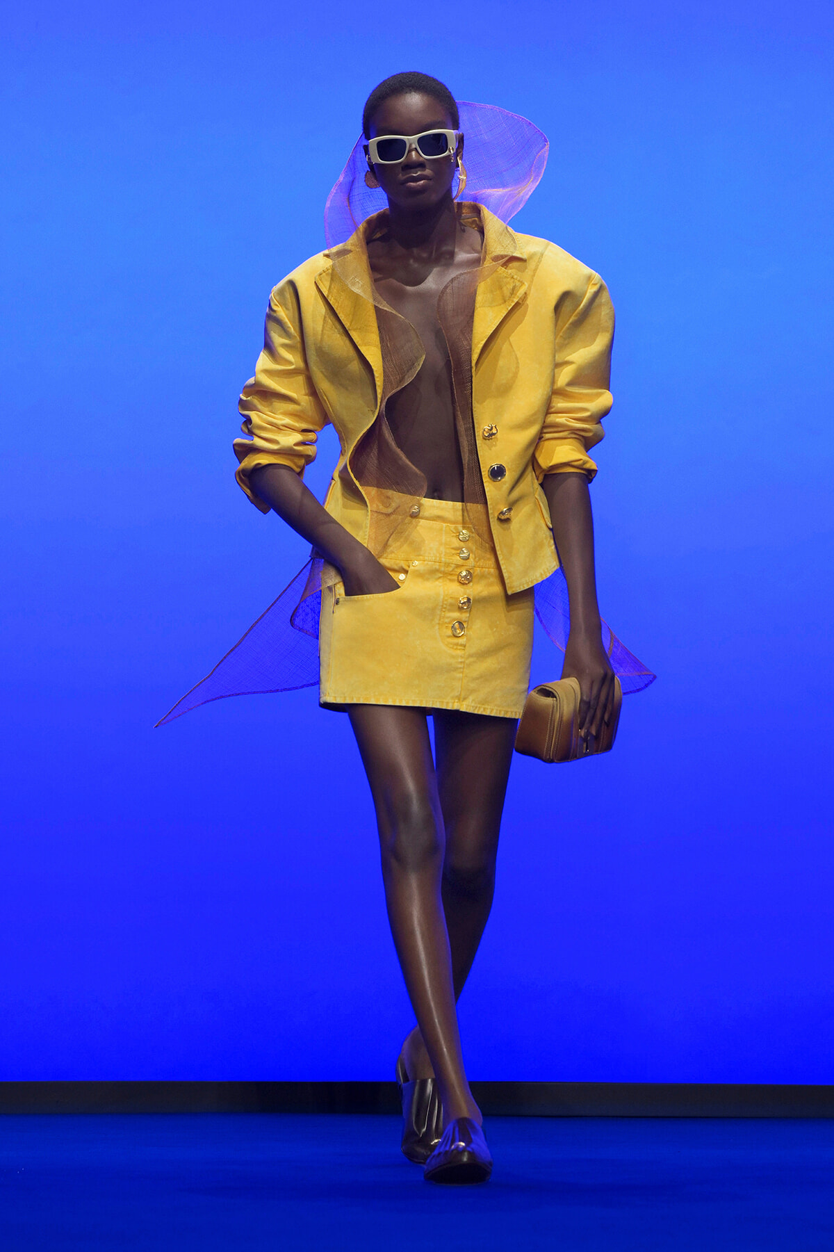 Model walks a blue runway in a bright yellow blazer and matching miniskirt, white sunglasses, sheer scarf, and a tan clutch.