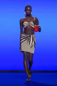 Model on a runway wearing a zebra-print bandeau top and knot-front mini skirt, with large earrings and a red handbag against a blue background.