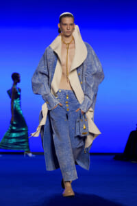 Model on the runway in an oversized light denim coat and matching jeans with a large cream collar and gold jewelry.