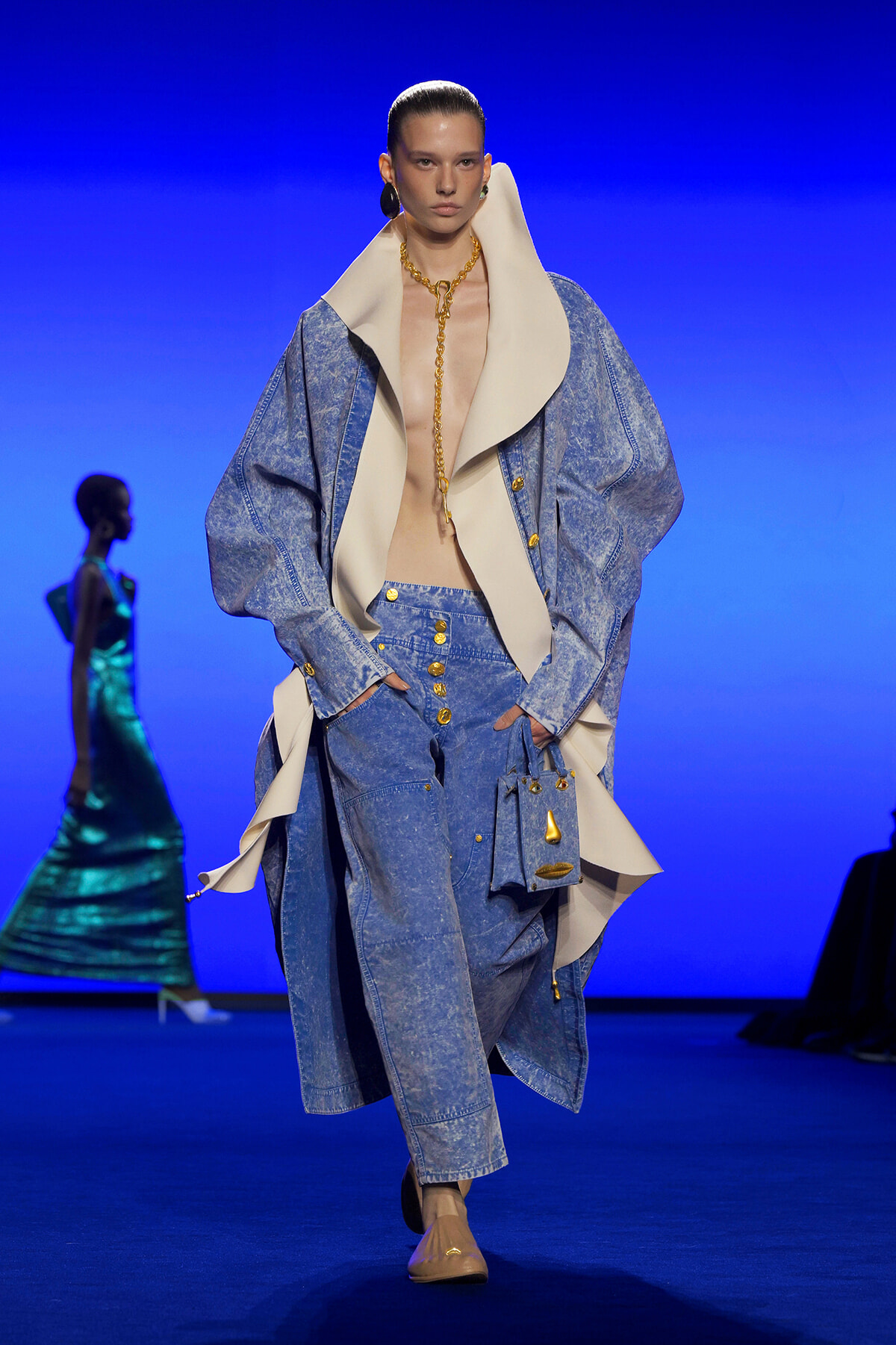 Model on the runway in an oversized light denim coat and matching jeans with a large cream collar and gold jewelry.