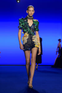 Model walking the runway in a short sequined dress with green and yellow floral appliqués and a large metallic neckpiece on a blue backdrop.