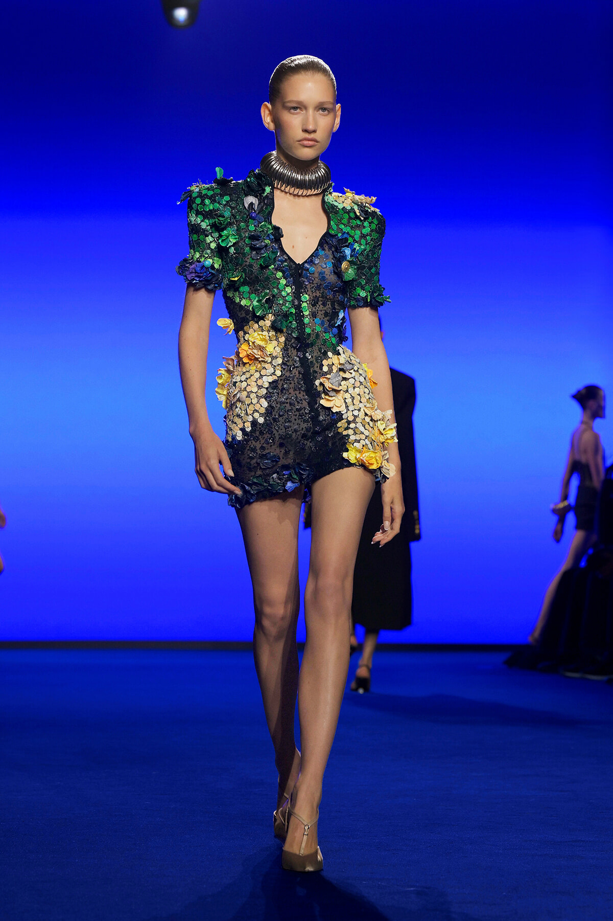 Model walking the runway in a short sequined dress with green and yellow floral appliqués and a large metallic neckpiece on a blue backdrop.