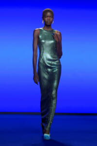 Model walking the runway in a metallic emerald-green sleeveless jumpsuit with sculpted seams, wearing gold earrings and a turquoise ring against a blue backdrop