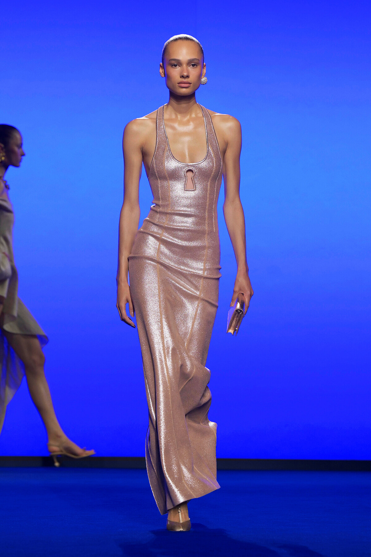 Model walking on a runway in a shimmering rose-gold halter gown with a keyhole neckline against a vivid blue backdrop