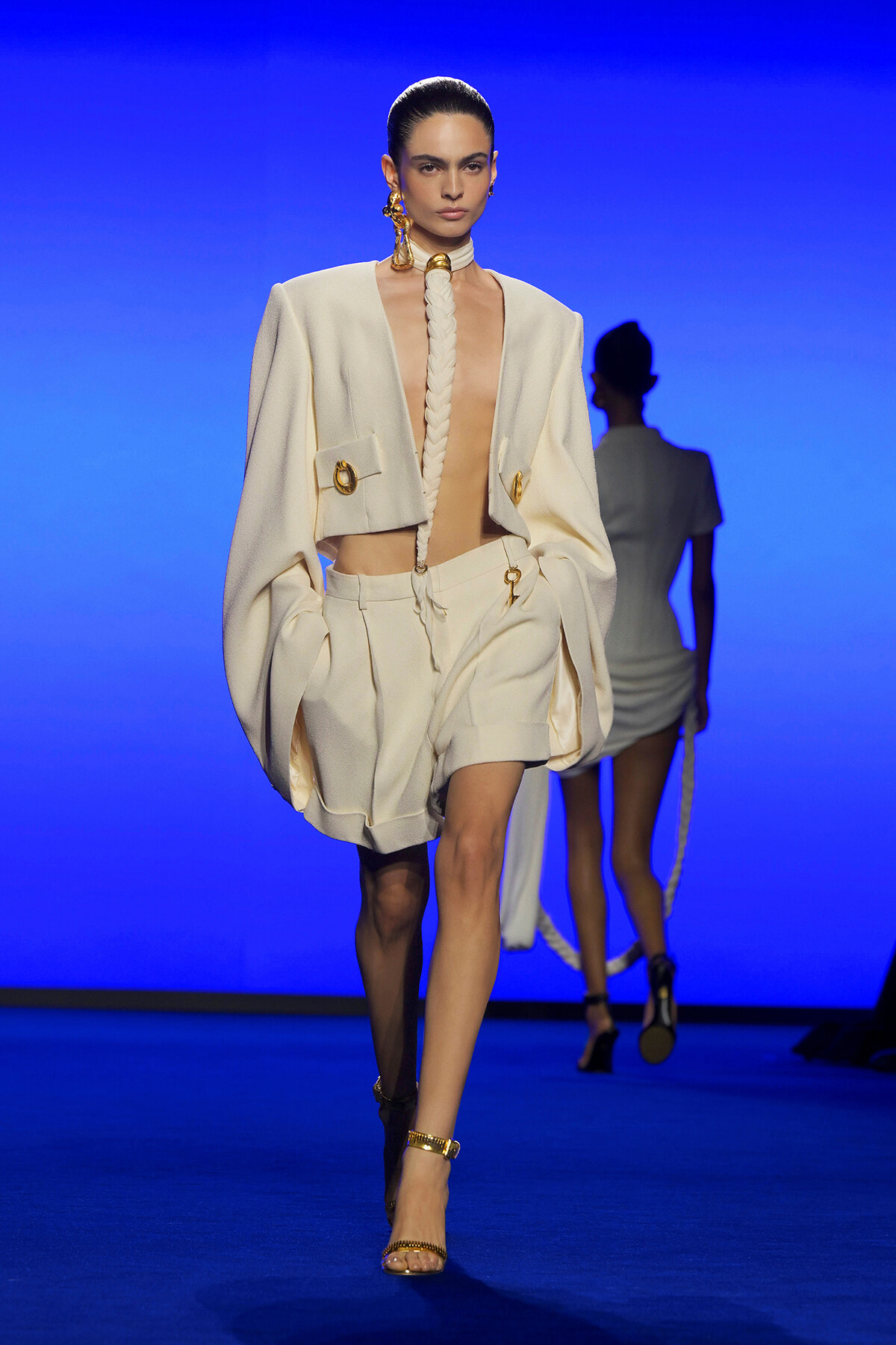 Model walks a runway in an ivory suit with oversized shoulders and a matching short bottom, paired with a braided rope necklace and gold jewelry against a blue backdrop.