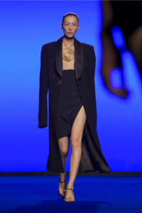 Model on a runway wearing a long black blazer over a strapless black dress with a high slit and a large gold sunburst necklace, against a blue backdrop.