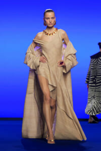 Model walks a runway in a beige textured, off-shoulder gown with oversized sleeves and cape, paired with bold red jewelry against a blue backdrop.
