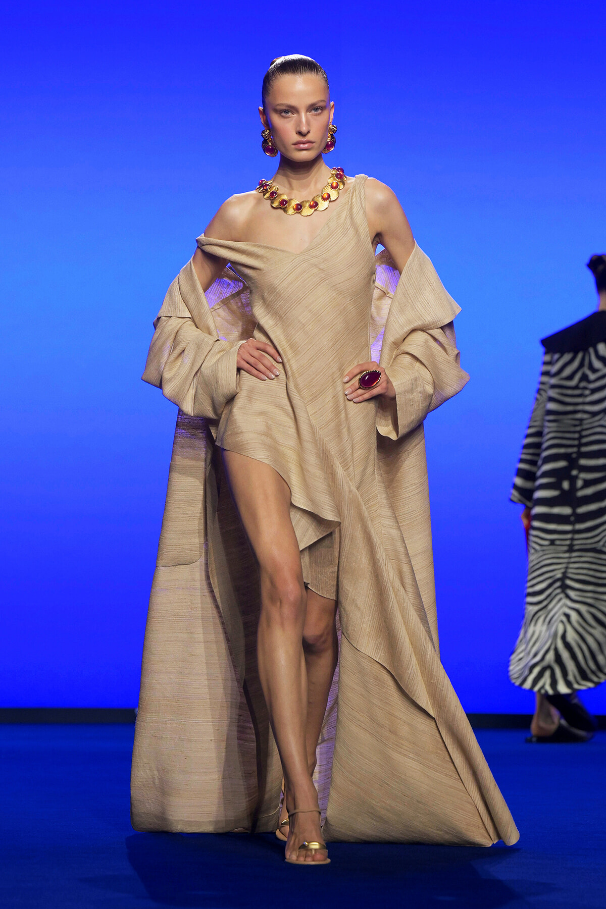 Model walks a runway in a beige textured, off-shoulder gown with oversized sleeves and cape, paired with bold red jewelry against a blue backdrop.