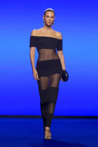 Model walking a runway in a black off-the-shoulder dress with sheer mesh panels and chunky hoop earrings against a blue backdrop.