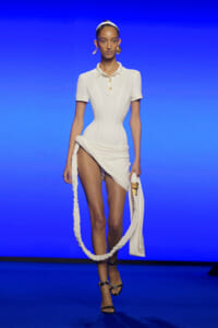 Model on a blue runway wearing a fitted white polo dress with a wrapped, asymmetric skirt and a long braided rope accessory held at her side.