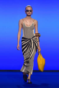 Model walking a runway in a cream-and-black striped dress with a front knot, gold chain straps, oversized yellow tote, and cat-eye sunglasses on a blue backdrop.