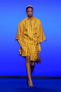Model walks a runway in a mustard-yellow plaid cape and matching shorts, accessorized with gold chains and a small brown handbag against a blue backdrop