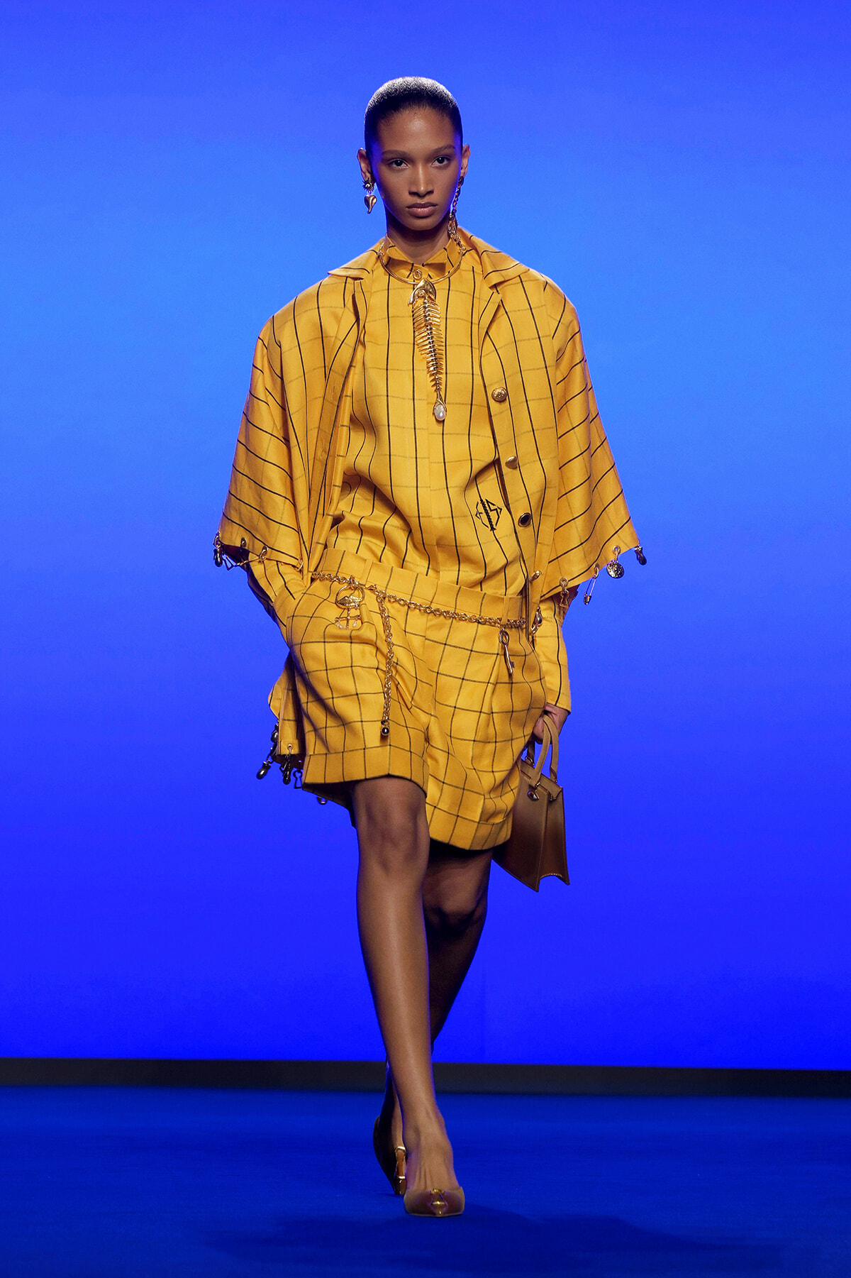 Model walks a runway in a mustard-yellow plaid cape and matching shorts, accessorized with gold chains and a small brown handbag against a blue backdrop
