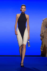 Model on a runway wearing a black halter top and cream draped skirt with a black front panel, carrying a small silver handbag.