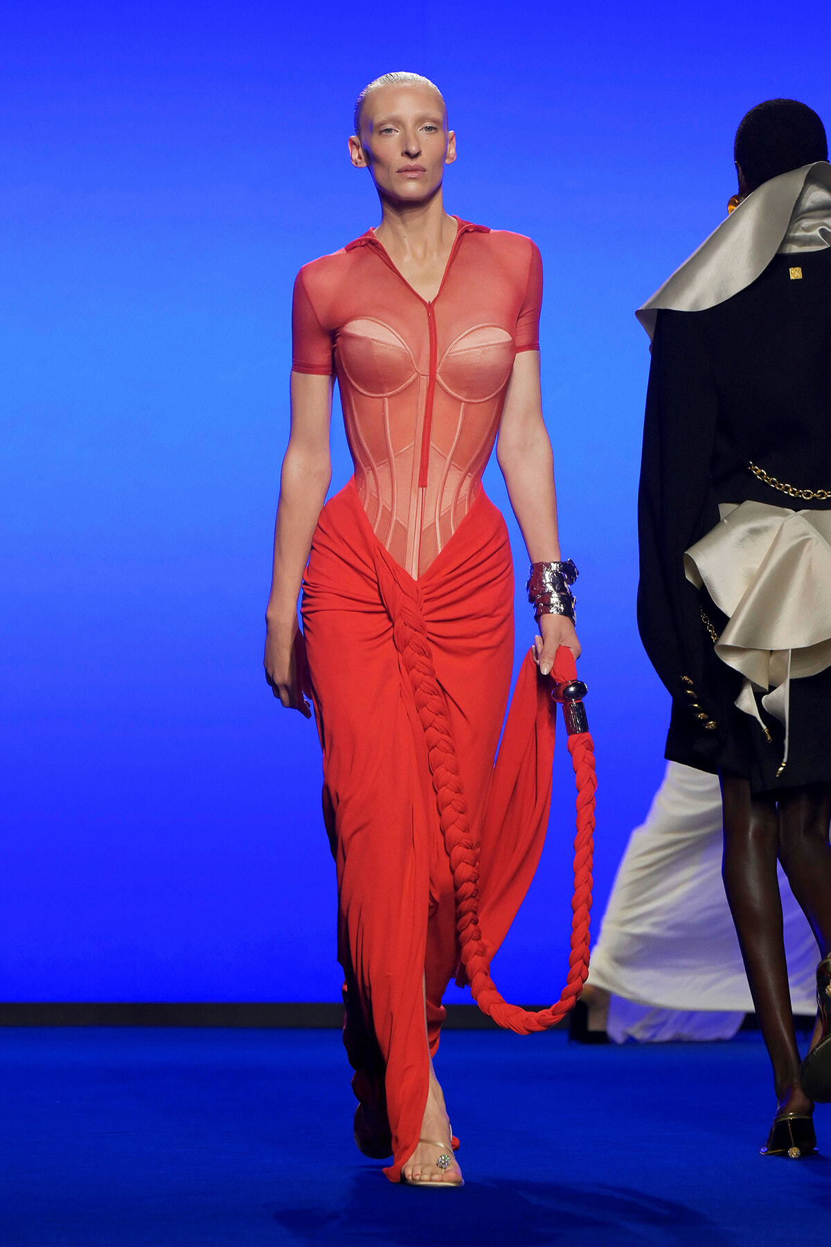 Model walks a red sheer corset top and draped red pants on a blue runway, holding a thick braided red rope accessory with a silver bracelet visible on one wrist.