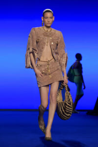Model on a runway wearing a tan denim jacket and mini skirt, gold jewelry, and zebra-print bag.