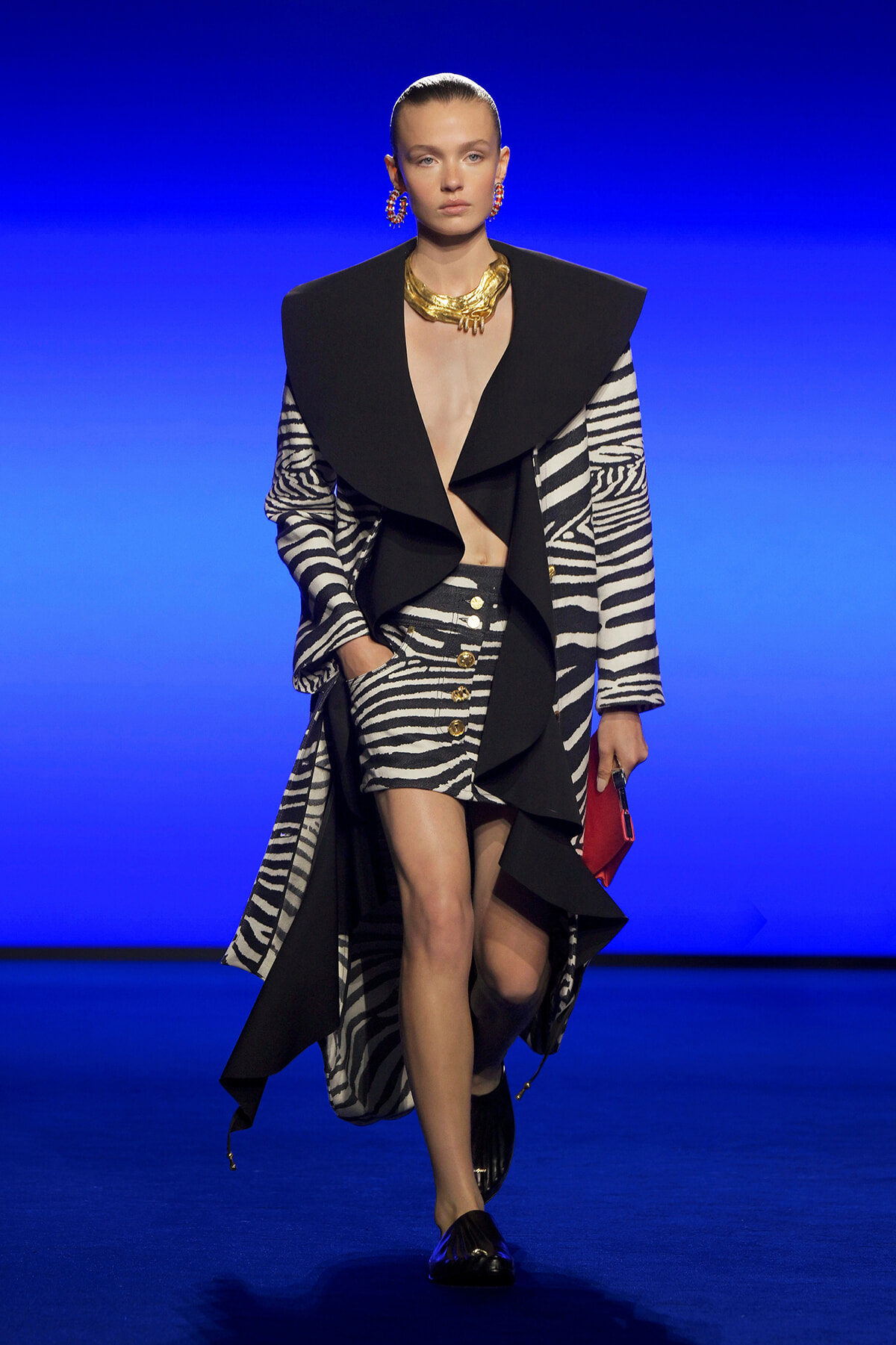 Model walks a fashion show wearing a black oversized-lapel coat over a zebra-print outfit with gold jewelry and a red clutch against a blue runway backdrop to convey high-fashion.”,