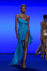 Model walks a blue-lit runway in an asymmetrical, draped turquoise gown with a high slit, wearing chunky silver jewelry and sandals on a blue stage.