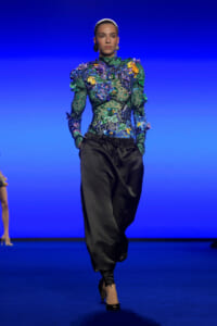 Model walks the runway in a green floral-embellished top and black satin harem pants against a blue backdrop.