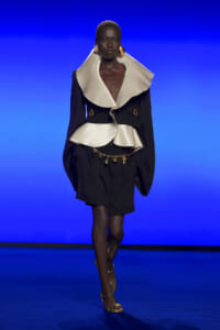 Model walks the runway in a black outfit with an oversized cream collar, gold belt rings, and large gold earrings against a blue backdrop.