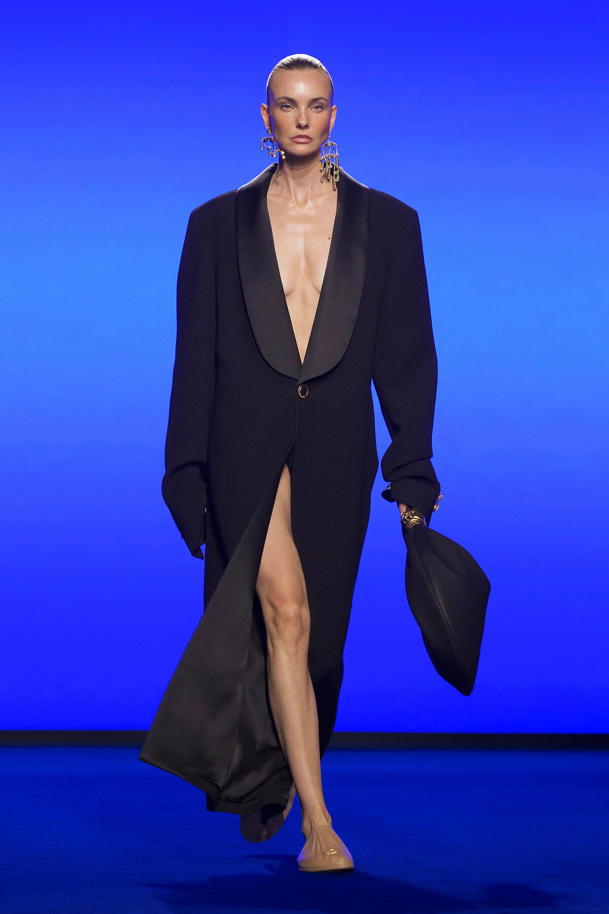 Female model walks a runway in a black oversized blazer with a plunging neckline and high slit, holding a black hat, wearing bold gold earrings against a blue backdrop.