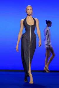 Model walking the runway in a sleeveless denim jumpsuit with a central gold zipper against a blue backdrop.