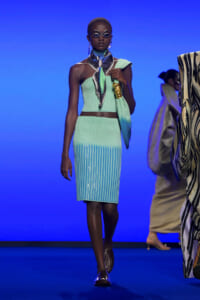 Model on a blue runway wearing a mint-green ribbed two-piece outfit with a scarf, metallic jewelry, and futuristic sunglasses.
