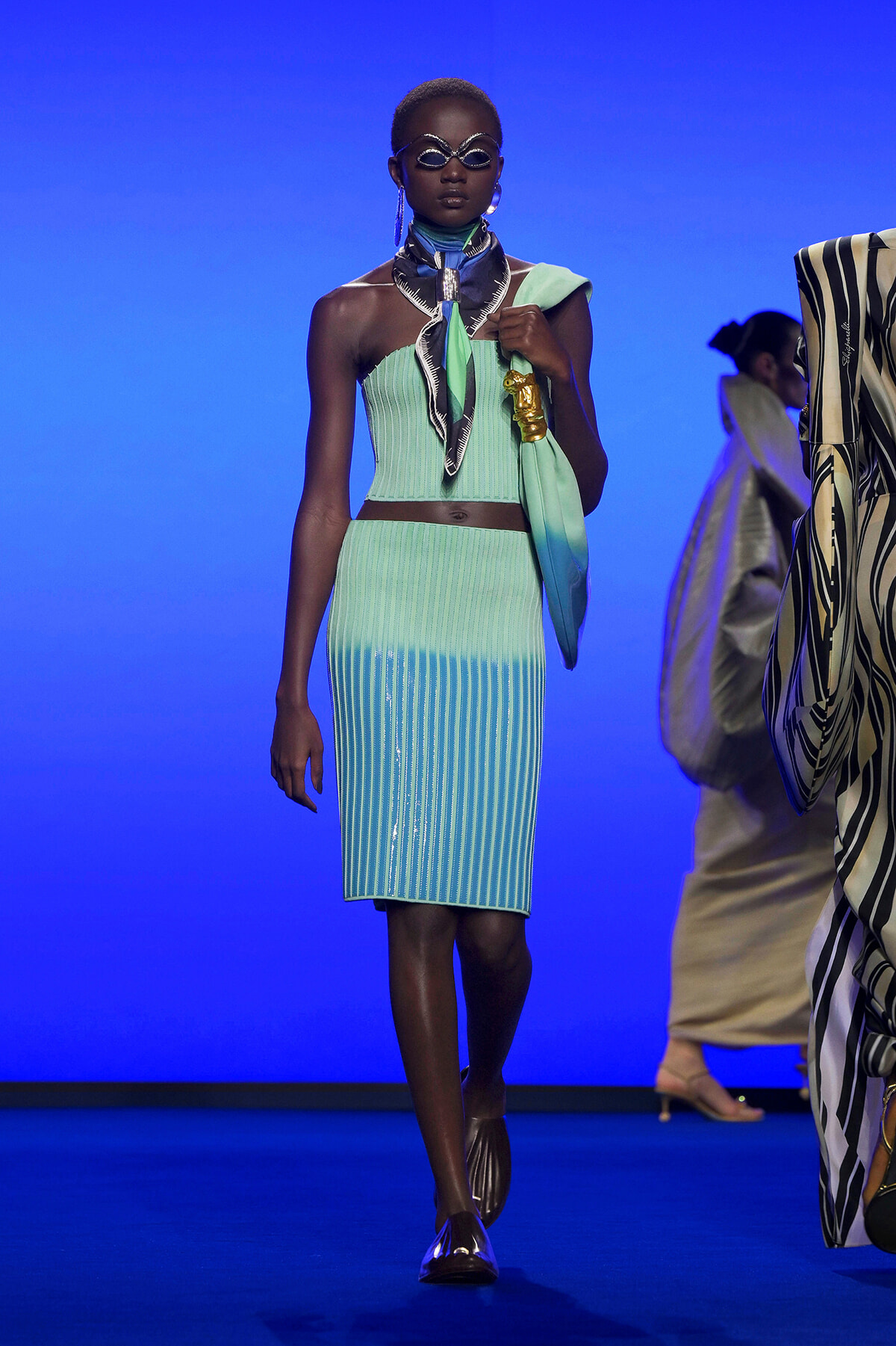 Model on a blue runway wearing a mint-green ribbed two-piece outfit with a scarf, metallic jewelry, and futuristic sunglasses.