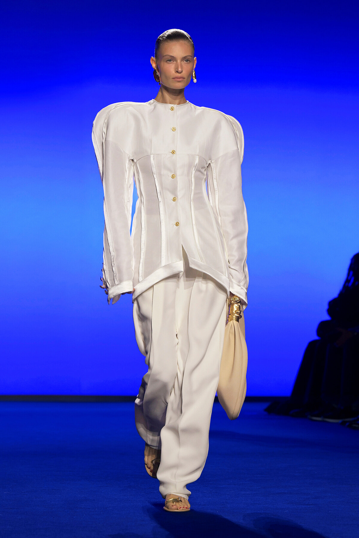 Model walking a runway in a white structured suit with oversized shoulders, gold buttons, and a cream bag against a blue backdrop.