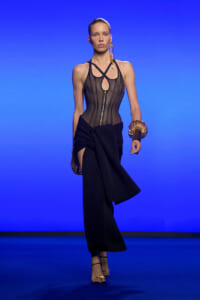 Model walking a fashion runway in a sheer black corset top with a front zipper and a black draped skirt, blue backdrop behind her, gold accessories on ears and wrist.