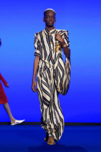 Model walking a fashion runway in a cream-and-black striped outfit with gold buttons and a gold bag, against a blue backdrop.