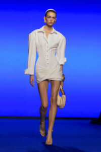 Model walks the runway in a white tailored shirt-dress with a corseted waist and gold buttons, blue backdrop behind her, holding a beige bag.