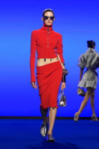 Model on a runway wearing a red cropped turtleneck sweater with a front zipper and a matching red pencil skirt, sunglasses, and metallic accessories against a blue backdrop.