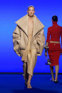 Model walks the runway in a large beige quilted coat with oversized collar, matching pants, and gold accessory against a blue backdrop